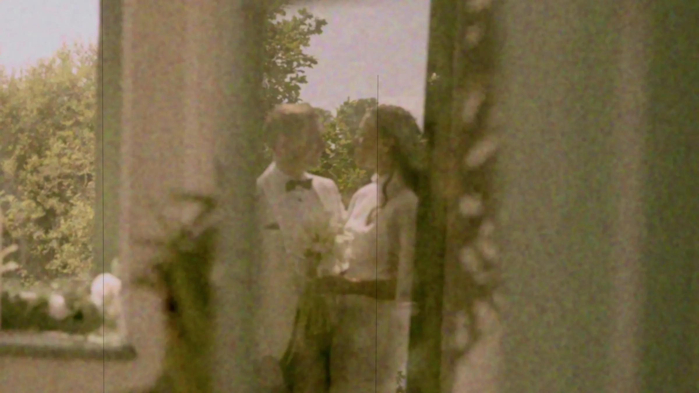 A grainy, Super 8-style film frame of a couple embracing beside a tall window, in soft colour with visible film texture.